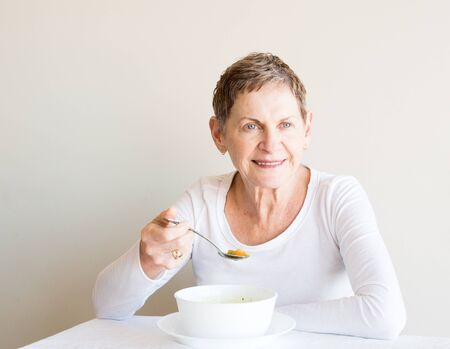 Older woman with short hair and white top smiling and eating soup from a white bowlの写真素材