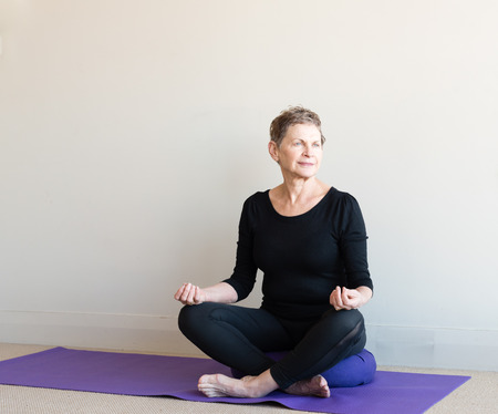 Older woman in black yoga clothing sitting cross legged on purple cushion in meditation posture with hands in guyan mudraの写真素材