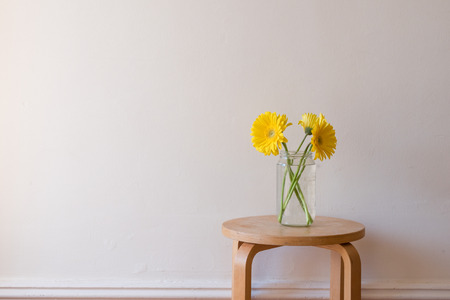 Yellow gerberas in a glass jar on small wooden table against vintage white wall (landscape orientation)の写真素材