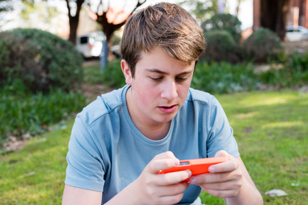 Teenage boy in blue t-shirt playing on his smart phone outdoors in park (selective focus)の写真素材
