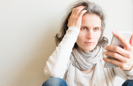 Middle aged woman with grey hair and cream top sitting on floor with hand on head using smart phone (selective focus and cropped)の写真素材