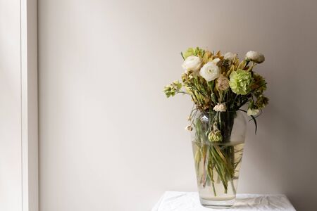 Wilting green and white ranunculus and carnations in large glass vase on small white table next to...