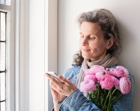 Middle aged woman with grey hair and denim shirt using smart phone, holding pink flowers and smiling