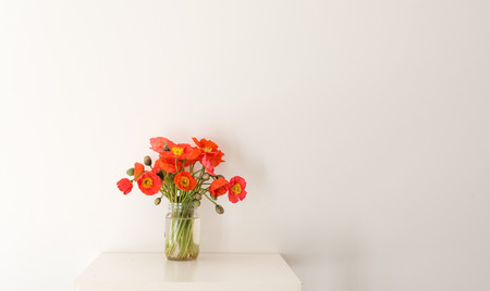 Red poppies in glass jar on white table against white wallの写真素材