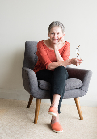 Older woman in orange top seated in armchair holding glasses and smilingの写真素材