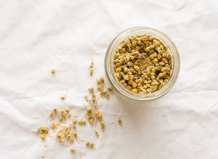Top down view of dried chamomile flowers for tea in glass jar and scattered on white tablecloth (selective focus)の写真素材