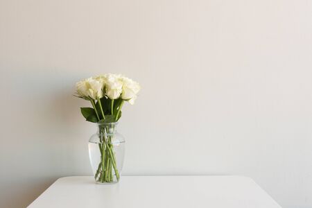 Cream roses in glass vase on white table against neutral wall