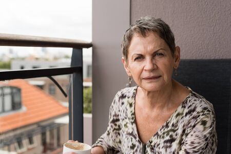 Older woman with coffee cup seated on balcony against urban background (selective focus)の写真素材