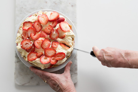 High angle view of older woman's hands holding knife and cutting strawberry and cream cheesecake on marble board (selective focus)の写真素材