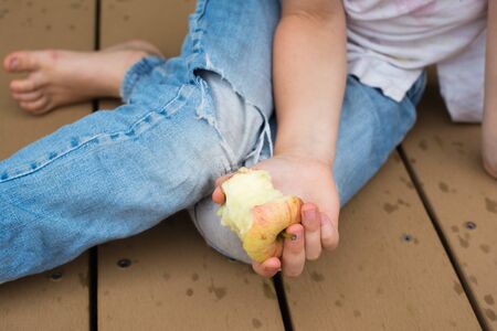Cropped view of child in denim jeans holding half eaten and sitting on floor (selective focus)