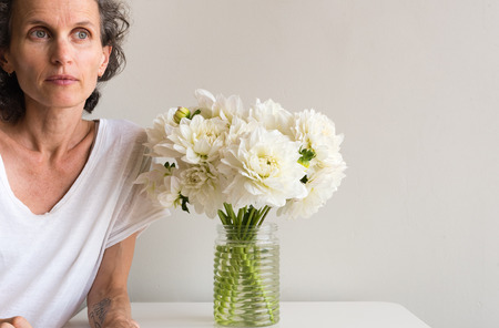 Middle aged woman with grey hair looking thoughtful leaning on table with white dahlias (cropped)の写真素材