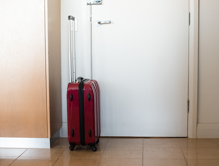 Small lightweight modern red suitcase by hotel room door (selective focus)の写真素材