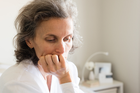 Close up head and shoulders view of tired middle aged woman resting head on hand wearing robe and seated on bed (selective focus)の写真素材