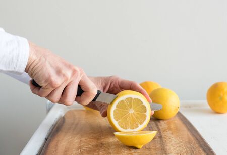 Close up of middle aged woman's hand holding knife and slicing lemon on wooden chopping board (selective focus)の写真素材