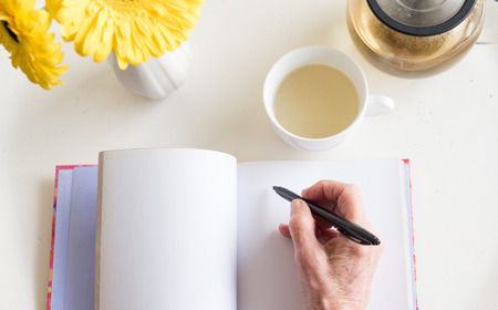 High angle view of older woman's hand holding black pen on blank journal with cup of chamomile tea and yellow flowers (selective focus)の写真素材