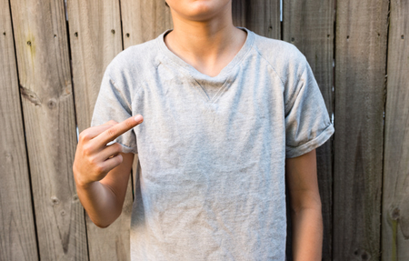 Cropped view of teenage boy in grey t-shirt making rude finger gesture against wooden fence (selective focus)の写真素材