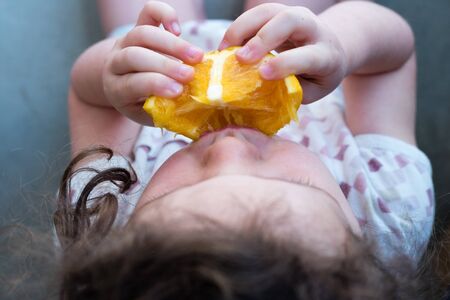 High angle view of little girl eating orange piece and sitting on floor (selective focus)の写真素材