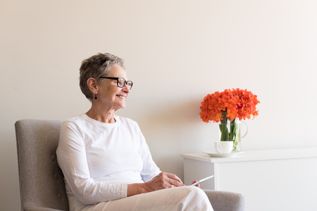 Older woman in black glasses with white clothing seated and using tablet next to orange clivia flowers (selective focus)の写真素材