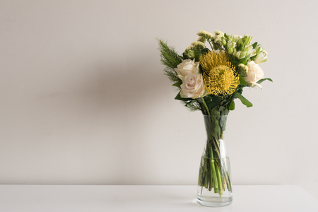 Close up of yellow and cream flowers in glass vase on white table against neutral wall background with copy spaceの写真素材