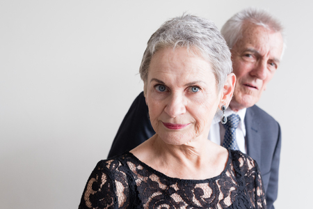 Beautiful older woman in black cocktail dress looking at camera with older man in suit in background (selective focus)の写真素材
