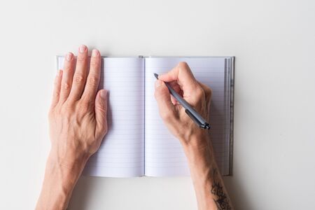 High angle view of woman's hands holding black pen above blank journal on white tableの写真素材