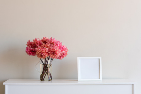 Bright coral pink dahlias in glass jug with blank square frame on white sideboard against neutral wall backgroundの写真素材