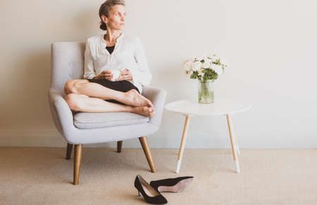 Middle aged woman relaxing with tea in armchair with shoes kicked off on floor - matte filter effect and selective focusの写真素材