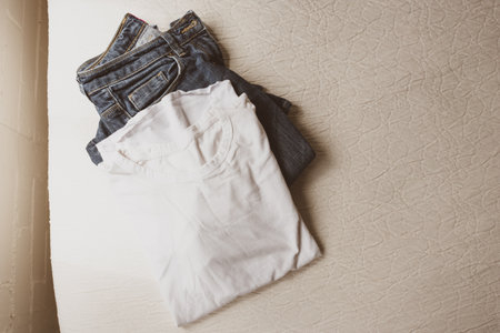 HIgh angle view of folded plain white t-shirt and blue jeans on edge of cream coloured bedding - warm matte filter effectの写真素材
