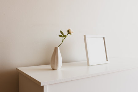 Close up of single rose in small white vase next to blank square picture frame on sideboard against neutral wall - warm matte filter effect and selective focusの写真素材