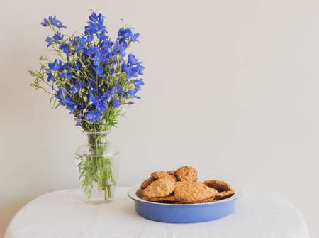 Blue delphinium flowers in glass vase on table with platter of homemade healthy cookies against...