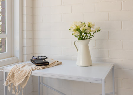 Closeup angled view of white freesias in jug on table with scarf, purse and glasses against painted...