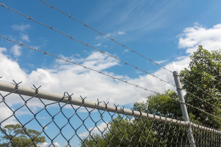 Close up of barbed wire and metal fence against blue skey with clouds and green trees - keep out...
