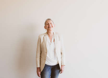 Three quarter length view of laughing older woman in cream jacket and jeans against neutral wall...