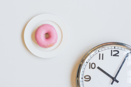 High angle view of pink donut on plate and clock face against white background - snacking conceptの写真素材