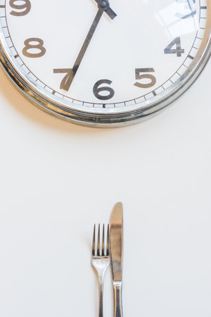 Vertical view of knife and fork with clock on white background - intermittent fasting concept (selective focus)の写真素材