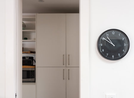 Black clock on white wall with view through doorway to tidy pantry utility room (selective focus)