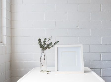 Closeup of blank square frame next to glass jar with eucalyptus leaves on white table