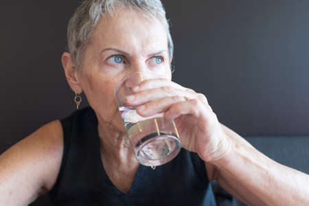Close up of beautiful older woman in black top drinking glass of water against dark background...
