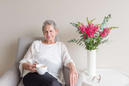 Older woman with short grey hair sitting in armchair drinking tea against neutral wall background...