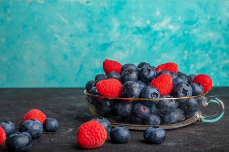 Fresh blueberries and strawberries in a glass bowl and on a dark table with turquoise backgroundの写真素材