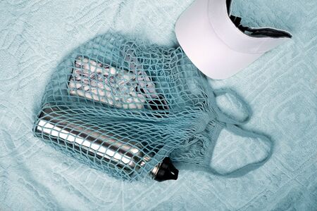 Bottle of water, mobile phone and smart watch in blue string mesh reusable bag and white visor on the beach towelの写真素材