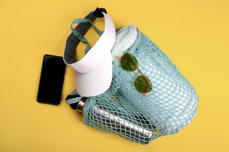 Bottle of water and beach towel in blue string mesh reusable bag with mobile phone, white visor and sun glasses on the yellow backgroundの写真素材