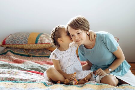 Mother and her small daughter are talking on the bed. The girl says something on moms ear.の写真素材