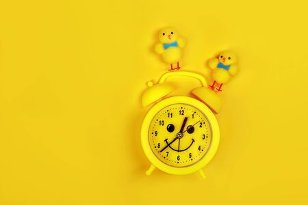 Retro yellow alarm clock with drawn smiling face and toys chicken and colored egg on yellow background. Easter concept.の写真素材