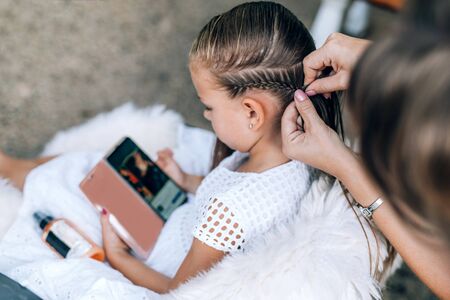 Mother does hair braid to her daughter, the girl is watching a cartoon on a mobile phone. Close up photo.の写真素材