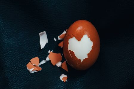 Cracked hard boiled brown egg with heart shaped shell on black leather background. Minimal Easter...