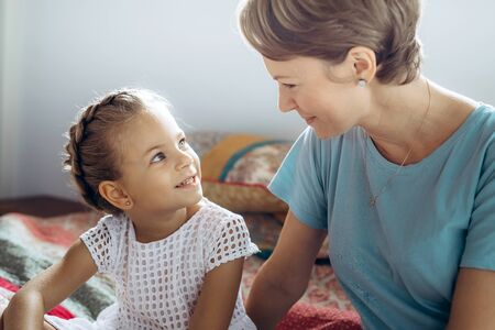 Mother and her small daughter are talking on the bed, looking at each other.