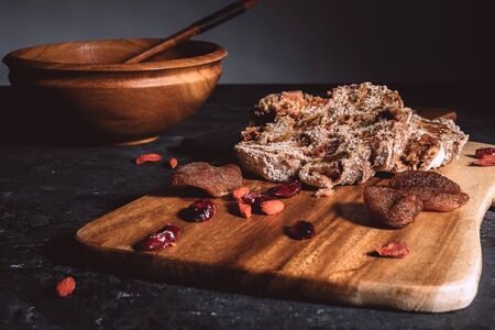 Homemade fruit bread on wooden chopping board with scattered dry berries and wooden bowl on...