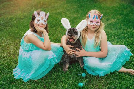 Cute Airedale Terrier dog in Easter bunny ears and little girls in bunny masks on Easter day sitting on grass in garden.の写真素材