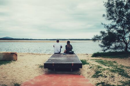 Young couple sitting together on a bench and looking at the river. Back view.の写真素材